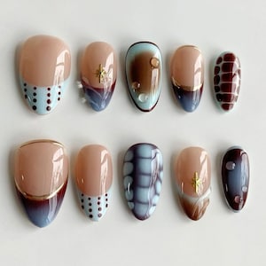 May include: A set of ten almond-shaped press-on nails. The nails feature a variety of designs, including light blue polka dots, gold accents, and abstract patterns in shades of brown, blue, and burgundy. Some nails have a nude base.
