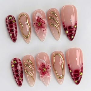 May include: A collection of ten artificial nails with diverse designs. Some nails have a pale pink base with gold detailing, while others display a deep red pattern. Two nails feature a 3D flower design, and some have gold studs.