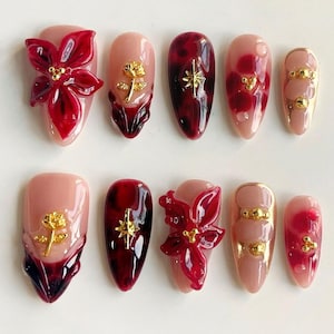 Peut inclure: Ensemble de dix faux ongles avec un motif rouge et or. Certains ongles présentent une fleur de poinsettia rouge avec des accents dorés, tandis que d'autres ont une étoile dorée ou une rose dorée.