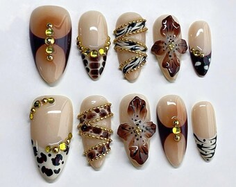 May include: A set of ten artificial nails with various designs. The nails feature a neutral base color with accents of brown, gold, and black. Designs include animal prints, floral patterns, and gold embellishments. The nails are oval-shaped.