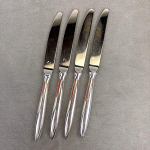 May include: Four silver knives with a polished, reflective finish. Each knife features a slender, curved blade and a rounded handle. The knives are aligned in a row, with the blades facing upwards. These knives are suitable for a formal dining occasion.