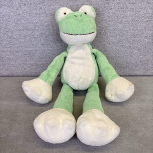 May include: A soft toy frog in green and white. The frog has large eyes, a smiling mouth, and long limbs. The toy is made of a soft material, suitable for children.