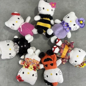 May include: A collection of white plush toys, each with a cat-like face and various outfits. The toys are dressed as a bee, in a kimono, and in other colorful outfits. Some have flower accents.