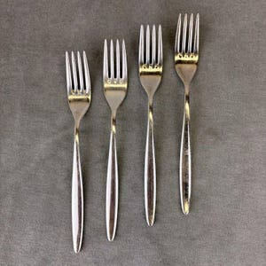 May include: Four silver forks of different sizes are displayed on a grey surface. The forks have a traditional design with tines and long handles. The forks are made of a shiny, reflective metal, probably stainless steel, and are suitable for dining.