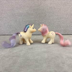 May include: Two cream-coloured pony figurines with long, brushable manes and tails. The left pony has a purple and blue mane with a shooting star symbol on its flank. The right pony features a pink and blue mane with three small star symbols.
