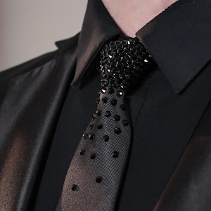 May include: A black necktie featuring a dense cluster of black faceted crystals at the knot, which gradually transition into a sparse, scattered pattern of individual black crystals extending down the length of the fabric against a black shirt.
