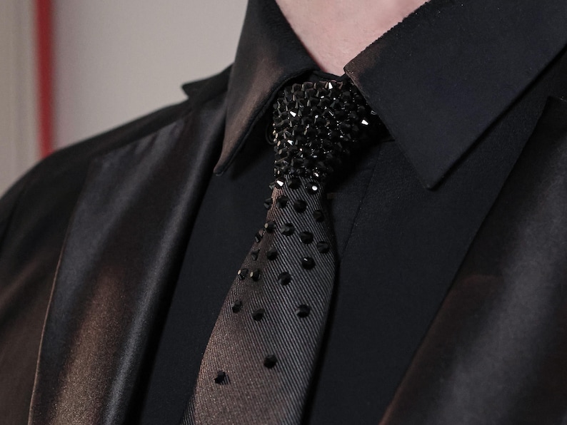 May include: A black necktie featuring a dense cluster of black faceted crystals at the knot, which gradually transition into a sparse, scattered pattern of individual black crystals extending down the length of the fabric against a black shirt.
