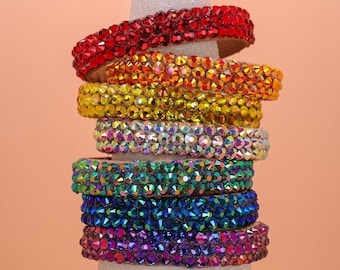 May include: A vertical stack of seven headbands, each covered in small, faceted, reflective crystals. The headbands are arranged in a rainbow color sequence, starting with red at the top, followed by orange, yellow, clear, green, blue, and purple.
