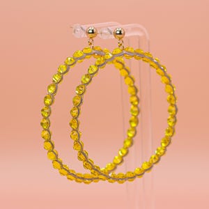 May include: A pair of large hoop earrings featuring a circular frame lined with small, faceted, bright yellow crystals. Each earring is attached to a polished gold-tone ball stud post, creating a vibrant and reflective accessory with a circular design.