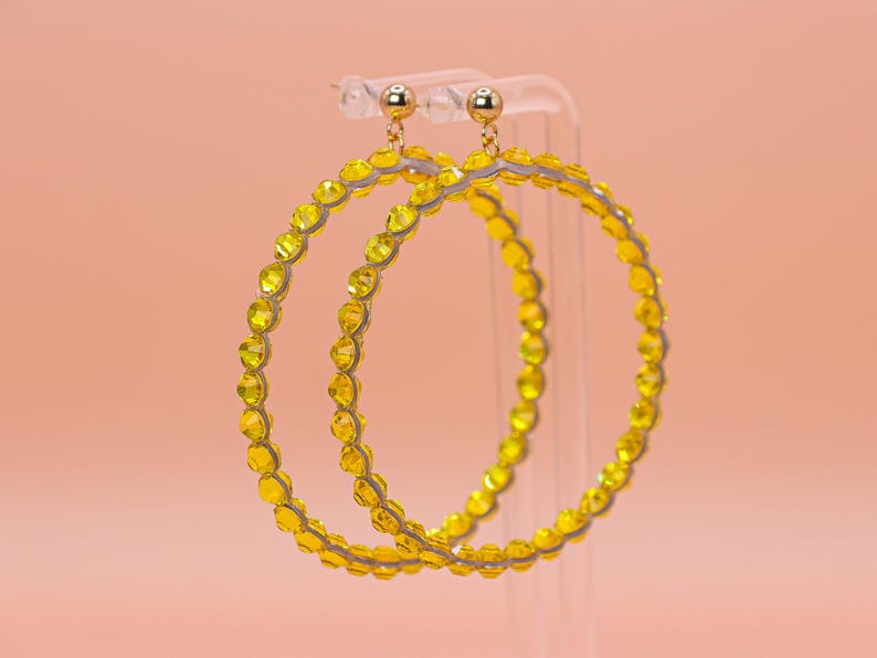 May include: A pair of large hoop earrings featuring a circular frame lined with small, faceted, bright yellow crystals. Each earring is attached to a polished gold-tone ball stud post, creating a vibrant and reflective accessory with a circular design.