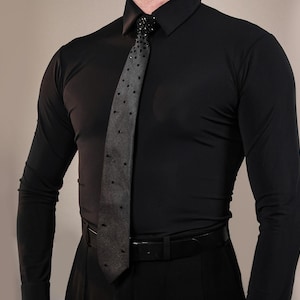 May include: A long-sleeved black button-down shirt paired with a dark grey necktie featuring a pattern of small black studs. The tie is secured at the collar with a decorative black beaded knot detail, and a black belt is worn around the waist.