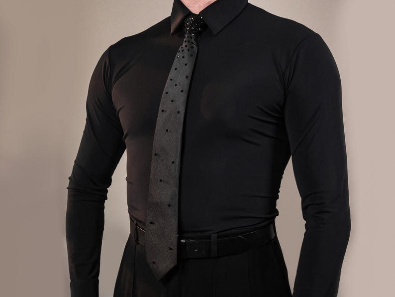 May include: A long-sleeved black button-down shirt paired with a dark grey necktie featuring a pattern of small black studs. The tie is secured at the collar with a decorative black beaded knot detail, and a black belt is worn around the waist.