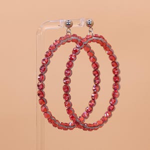 May include: A pair of circular hoop earrings featuring a continuous row of faceted red crystals set in a metallic frame. The earrings are suspended from small silver-toned ball studs, creating a bright and reflective appearance with a uniform design.