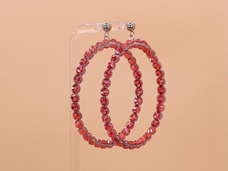 May include: A pair of circular hoop earrings featuring a continuous row of faceted red crystals set in a metallic frame. The earrings are suspended from small silver-toned ball studs, creating a bright and reflective appearance with a uniform design.