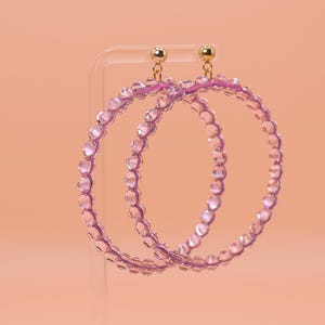May include: A pair of large hoop earrings featuring translucent light purple beads arranged in a circular pattern. The earrings are attached to small gold-toned metal ball studs at the top, providing a secure connection for the circular beaded hoops.