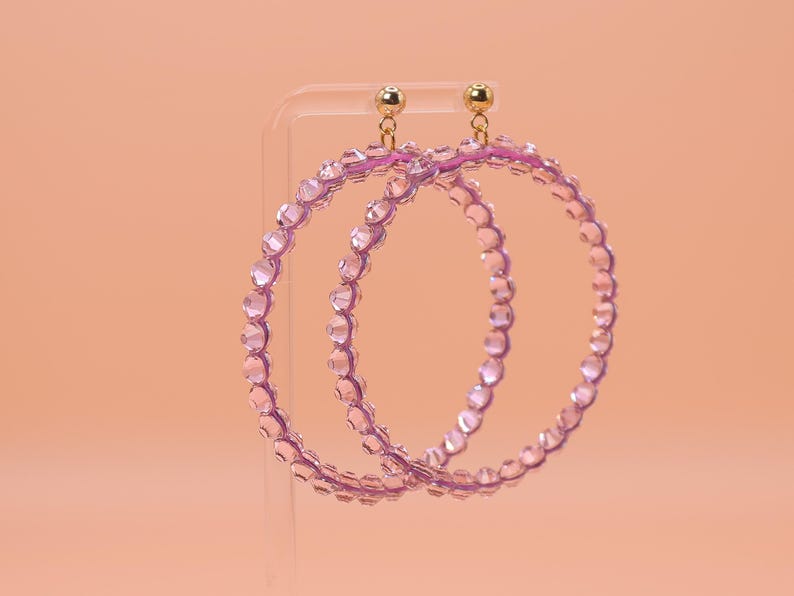 May include: A pair of large hoop earrings featuring translucent light purple beads arranged in a circular pattern. The earrings are attached to small gold-toned metal ball studs at the top, providing a secure connection for the circular beaded hoops.