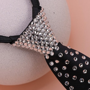 May include: A black necktie featuring a dense cluster of clear, reflective rhinestones covering the knot area, with additional individual rhinestones scattered across the length of the black fabric, attached to a thin black elastic neck band.
