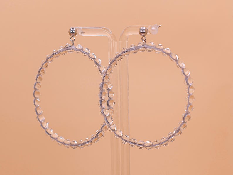May include: A pair of large hoop earrings featuring a clear, faceted bead design. The circular hoops are constructed from a series of transparent, light-reflecting beads strung together, finished with silver-toned metal posts and earring backs.