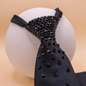 May include: A black tie with a beaded top section and scattered black gems. The tie is set against a neutral background, highlighting its dark colour and texture. The tie's design suggests formal wear.