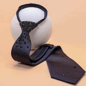 May include: A black necktie featuring a zipper mechanism and a series of small, dark, pointed studs embedded along the front surface. The tie has a textured fabric finish and includes a loop at the top for securing around the neck area.