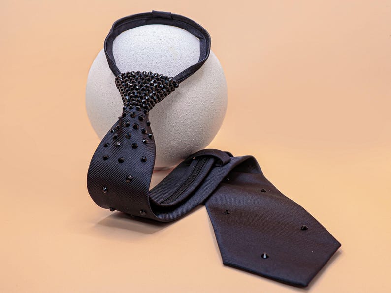 May include: A black necktie featuring a zipper mechanism and a series of small, dark, pointed studs embedded along the front surface. The tie has a textured fabric finish and includes a loop at the top for securing around the neck area.