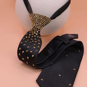 May include: A black necktie featuring a textured fabric surface adorned with numerous small, metallic gold-toned studs. The knot area is densely covered in these gold studs, which gradually become more sparse along the length of the tie.