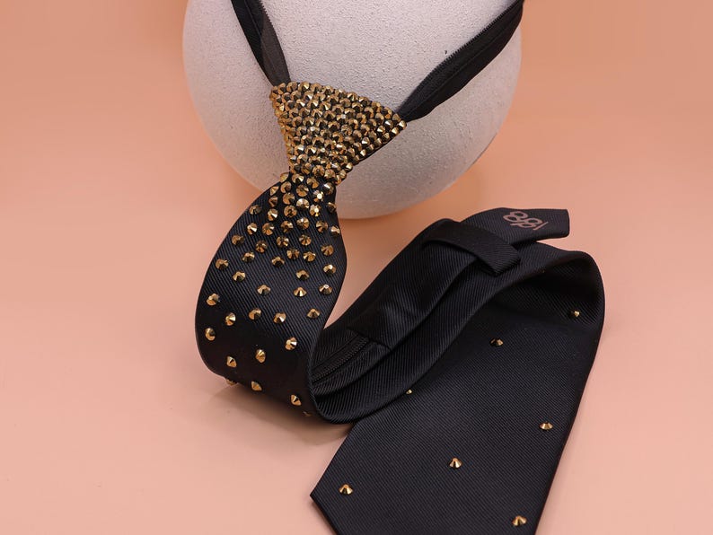 May include: A black necktie featuring a textured fabric surface adorned with numerous small, metallic gold-toned studs. The knot area is densely covered in these gold studs, which gradually become more sparse along the length of the tie.