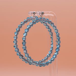 May include: A pair of hoop earrings featuring a circular frame adorned with small, faceted light blue beads. The earrings are suspended from silver-toned metal posts and are designed with a continuous row of translucent, sparkling blue crystals.
