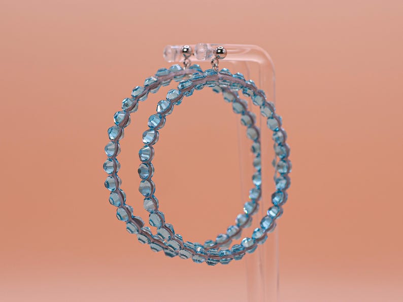 May include: A pair of hoop earrings featuring a circular frame adorned with small, faceted light blue beads. The earrings are suspended from silver-toned metal posts and are designed with a continuous row of translucent, sparkling blue crystals.