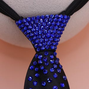 May include: A black necktie featuring a dense arrangement of faceted royal blue crystals covering the knot, with the crystals gradually thinning out as they extend downward along the length of the black fabric tie surface.