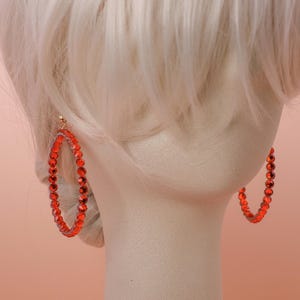 May include: A pair of large hoop earrings featuring a circular frame densely set with vibrant red faceted crystals. The earrings are displayed on a neutral-toned mannequin head with light-colored hair, highlighting the bright red color and the reflective quality of the individual stones arranged along the entire circumference of each hoop.