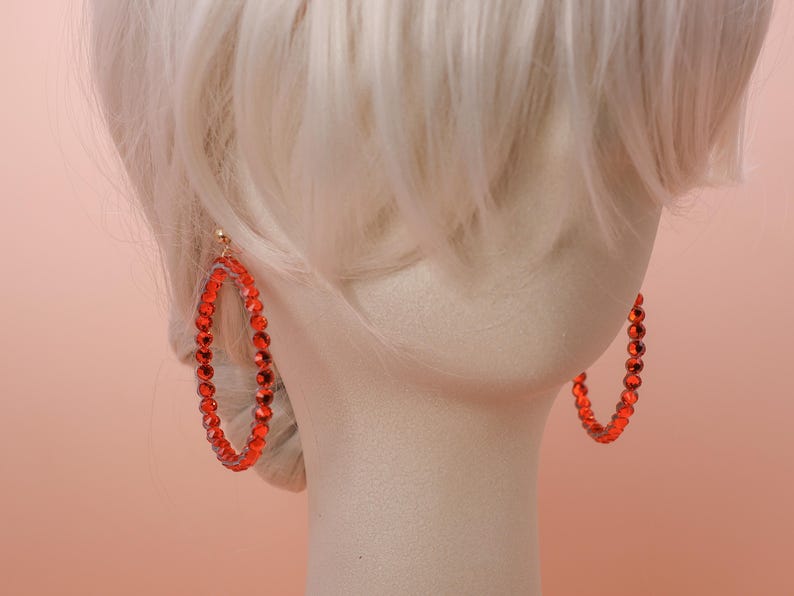 May include: A pair of large hoop earrings featuring a circular frame densely set with vibrant red faceted crystals. The earrings are displayed on a neutral-toned mannequin head with light-colored hair, highlighting the bright red color and the reflective quality of the individual stones arranged along the entire circumference of each hoop.