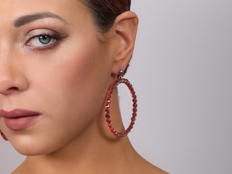 May include: A large circular hoop earring featuring a continuous row of small, faceted red beads set on a silver-toned frame. The earring attaches to the earlobe with a small silver-toned stud mechanism, creating a bold and vibrant accessory.