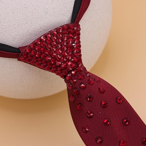 May include: A burgundy tie with a triangular knot fully encrusted with red rhinestones. The tie's lower portion features scattered red rhinestones. The tie is displayed against a neutral background, highlighting its sparkling details.