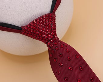 May include: A burgundy tie with a triangular knot fully encrusted with red rhinestones. The tie's lower portion features scattered red rhinestones. The tie is displayed against a neutral background, highlighting its sparkling details.