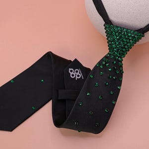 May include: A black necktie featuring a dense cluster of green crystals at the knot that gradually disperses into scattered individual green crystals along the length of the fabric. The back of the tie displays a white logo with the letters dp.