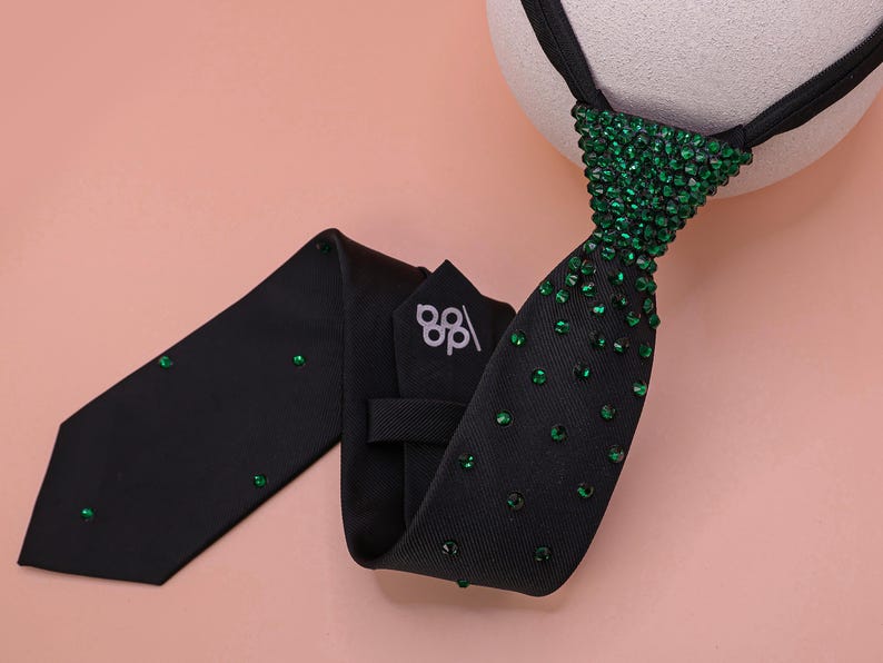 May include: A black necktie featuring a dense cluster of green crystals at the knot that gradually disperses into scattered individual green crystals along the length of the fabric. The back of the tie displays a white logo with the letters dp.