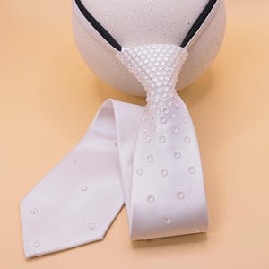 May include: A white tie with a textured knot and a dotted pattern on the tie's fabric. The tie is draped over a white spherical object with a black band. The background is a solid light orange color.