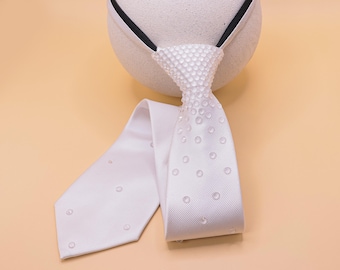 May include: A white tie with a textured knot and a dotted pattern on the tie's fabric. The tie is draped over a white spherical object with a black band. The background is a solid light orange color.
