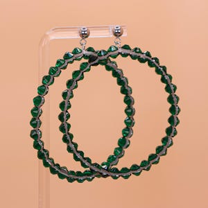May include: A pair of large hoop earrings featuring a circular frame covered in faceted emerald green beads. The earrings have silver-toned metal stud posts with small spherical accents at the top, connecting to the beaded hoops via thin metal rings.