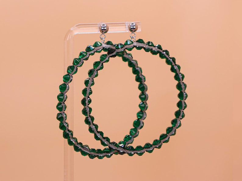 May include: A pair of large hoop earrings featuring a circular frame covered in faceted emerald green beads. The earrings have silver-toned metal stud posts with small spherical accents at the top, connecting to the beaded hoops via thin metal rings.