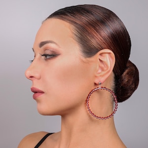 May include: A large circular hoop earring featuring a series of small, translucent, deep red beads strung together to form the hoop structure. The earring is attached to a small, silver-toned metal stud component that secures it to the earlobe.
