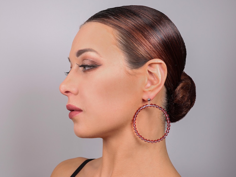 May include: A large circular hoop earring featuring a series of small, translucent, deep red beads strung together to form the hoop structure. The earring is attached to a small, silver-toned metal stud component that secures it to the earlobe.