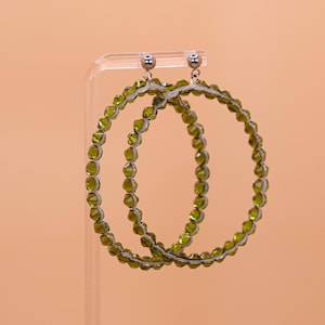 May include: A pair of large hoop earrings featuring a circular frame lined with small, faceted olive green beads. The earrings are attached to silver-toned metal stud posts with a polished finish, creating a circular design with a textured appearance.