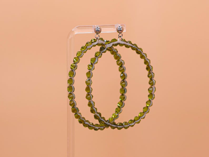 May include: A pair of large hoop earrings featuring a circular frame lined with small, faceted olive green beads. The earrings are attached to silver-toned metal stud posts with a polished finish, creating a circular design with a textured appearance.