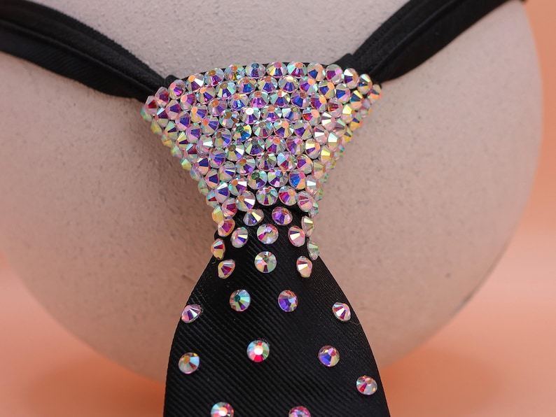 May include: A black necktie featuring a dense cluster of iridescent, multi-colored rhinestones at the knot that gradually transition into a sparse, scattered pattern of individual crystals extending down the length of the fabric.