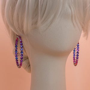 May include: A pair of large hoop earrings featuring a circular design encrusted with small, faceted crystals in a vibrant gradient of blue, purple, and pink hues. The crystals are set in a metallic frame, creating a reflective and colorful appearance.
