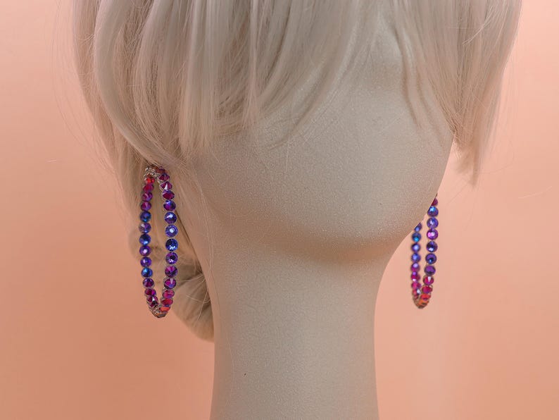 May include: A pair of large hoop earrings featuring a circular design encrusted with small, faceted crystals in a vibrant gradient of blue, purple, and pink hues. The crystals are set in a metallic frame, creating a reflective and colorful appearance.