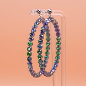 May include: A pair of circular hoop earrings featuring a continuous row of faceted, iridescent crystals that shift in color between deep purple, blue, and vibrant green. The stones are set in a metallic frame with small silver-toned stud posts.