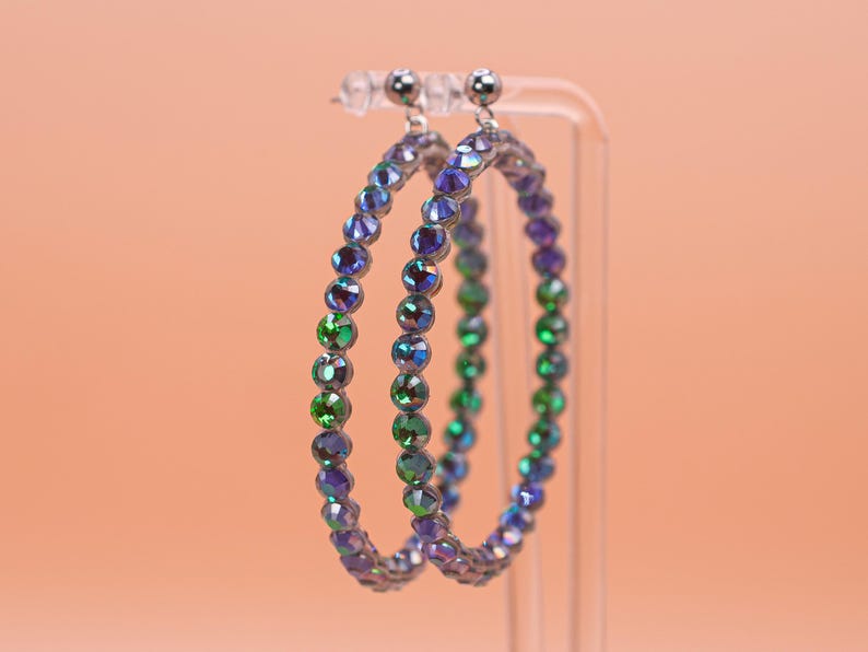 May include: A pair of circular hoop earrings featuring a continuous row of faceted, iridescent crystals that shift in color between deep purple, blue, and vibrant green. The stones are set in a metallic frame with small silver-toned stud posts.